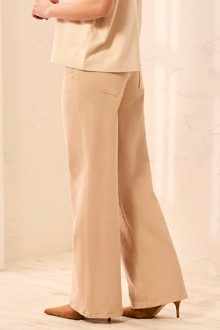 A woman shown from the back wearing tan wide leg shimmery pants and cream t-shirt standing in front of a beige background. 
