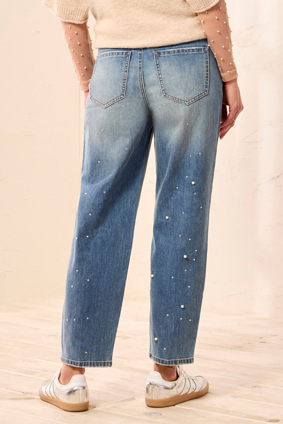 A woman shown from the back wearing pearl embellished barrel style washed jeans and a cream sweater standing in front of a beige background. 