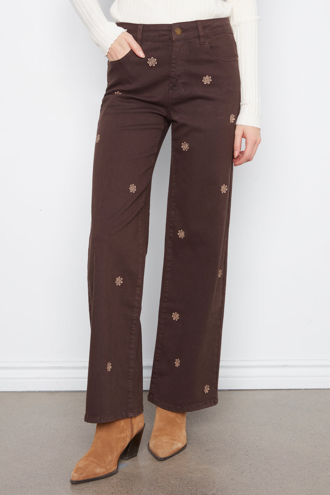 A woman wearing brown wide leg jeans with daisy embroidery standing in front of a grey wall. 