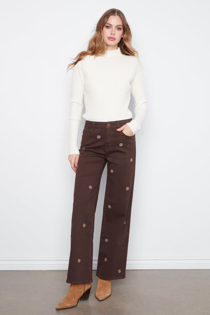A woman wearing brown wide leg jeans with daisy embroidery standing in front of a grey wall. 