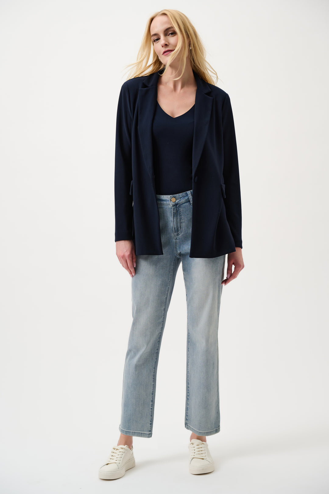 Woman wearing a black blazer, black top, light blue jeans, and white sneakers on a white background