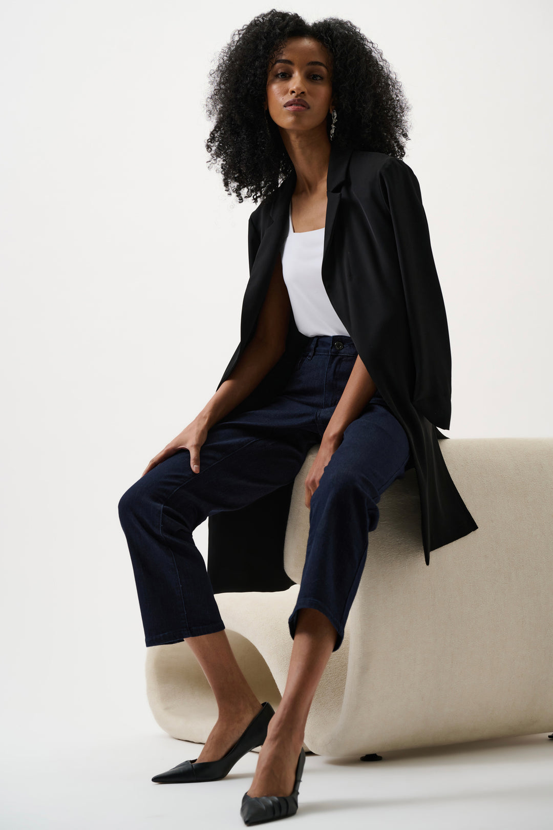Woman in black coat and navy jeans sitting on a white couch against a white background