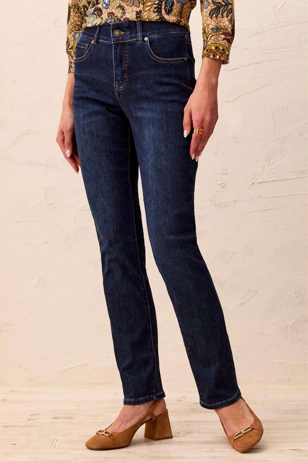 A person wearing dark blue slim-fit jeans with a patterned blouse and tan block-heel mules, standing against a light beige textured wall.