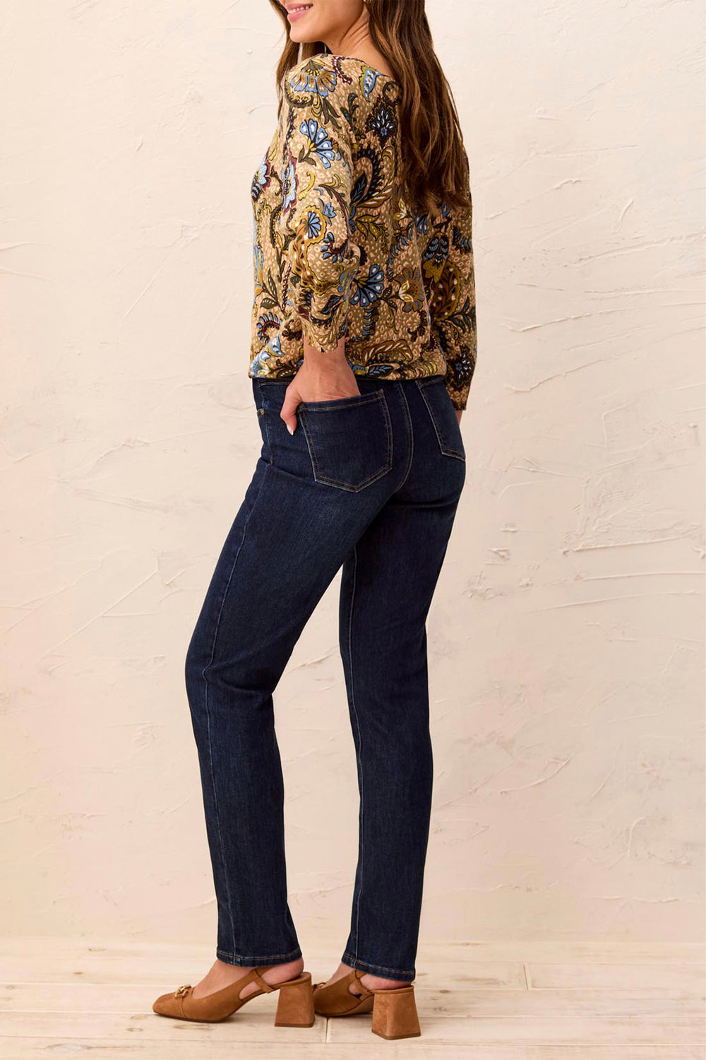 Woman wearing a floral blouse and dark jeans against a beige wall.