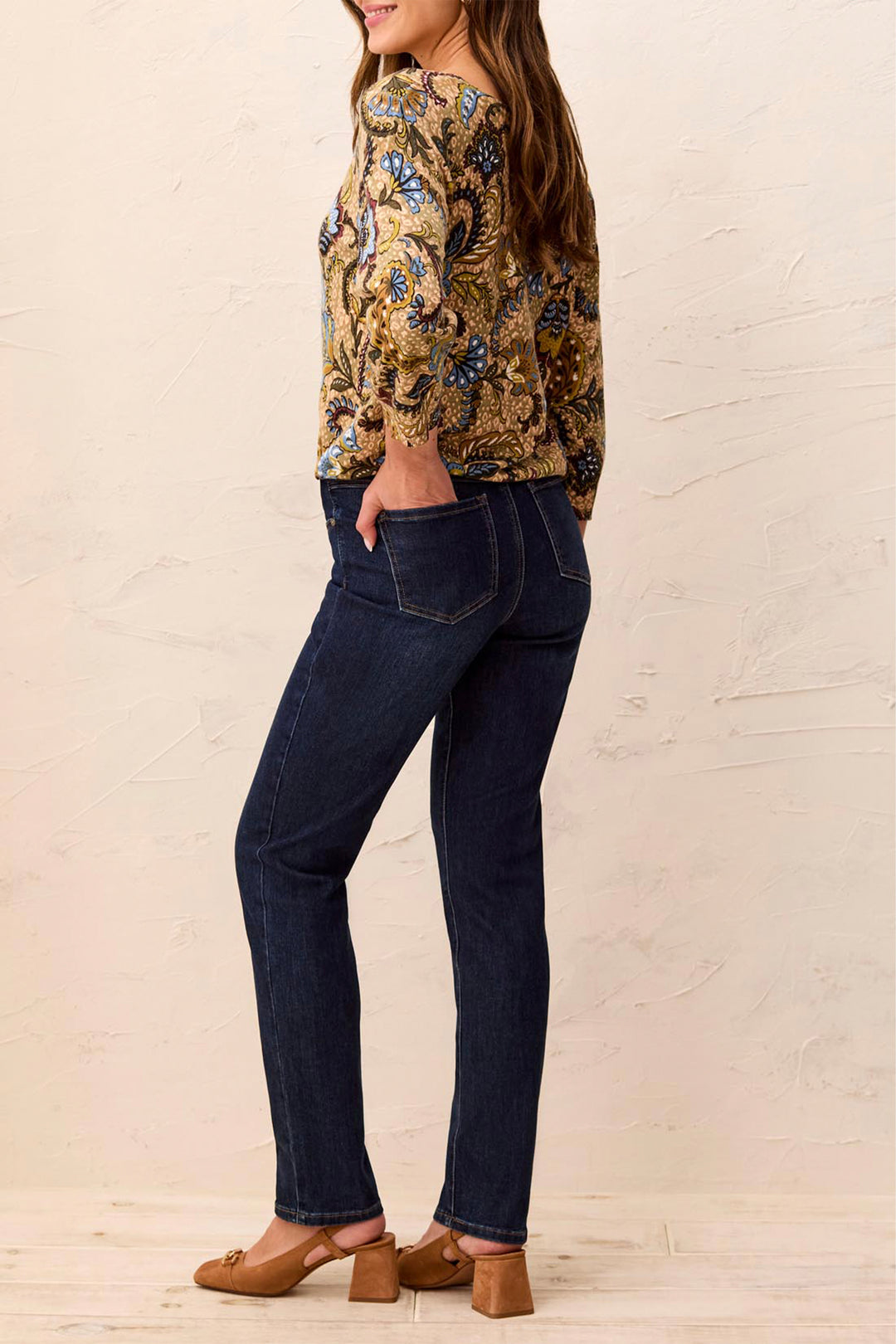Woman wearing a floral blouse and dark jeans against a beige wall.