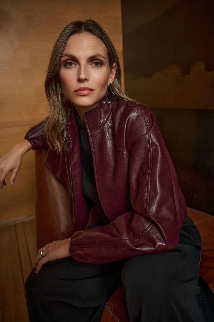 Woman wearing a burgundy pleather bomber jacket sitting on a wooden bench.