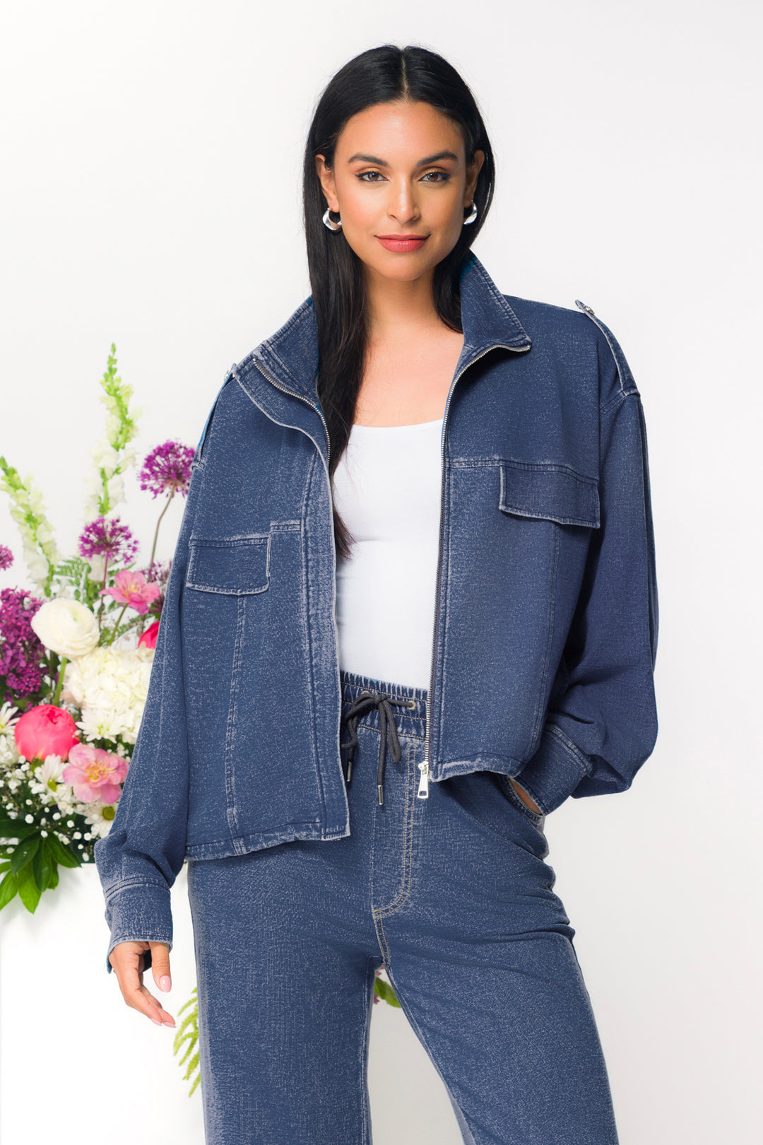 Woman wearing a denim sweater jacket and jeans with a white background and flowers.