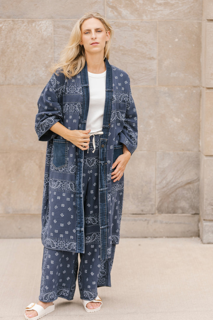 Woman wearing a blue long open cardigan standing against a stone wall.