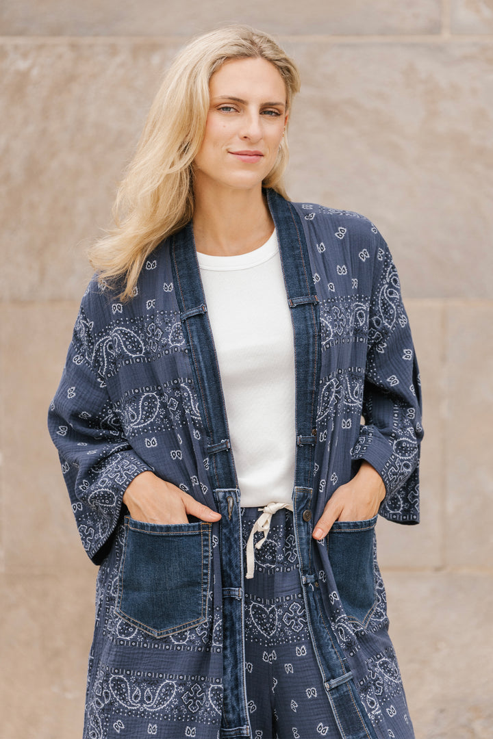 Woman wearing a blue long open cardigan robe over a white shirt against a beige wall.
