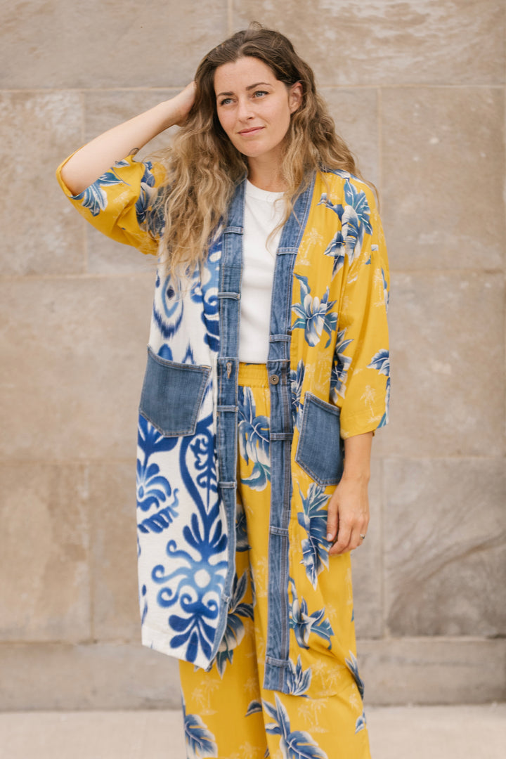 Woman wearing a yellow floral kimono over a denim jacket against a stone wall.
