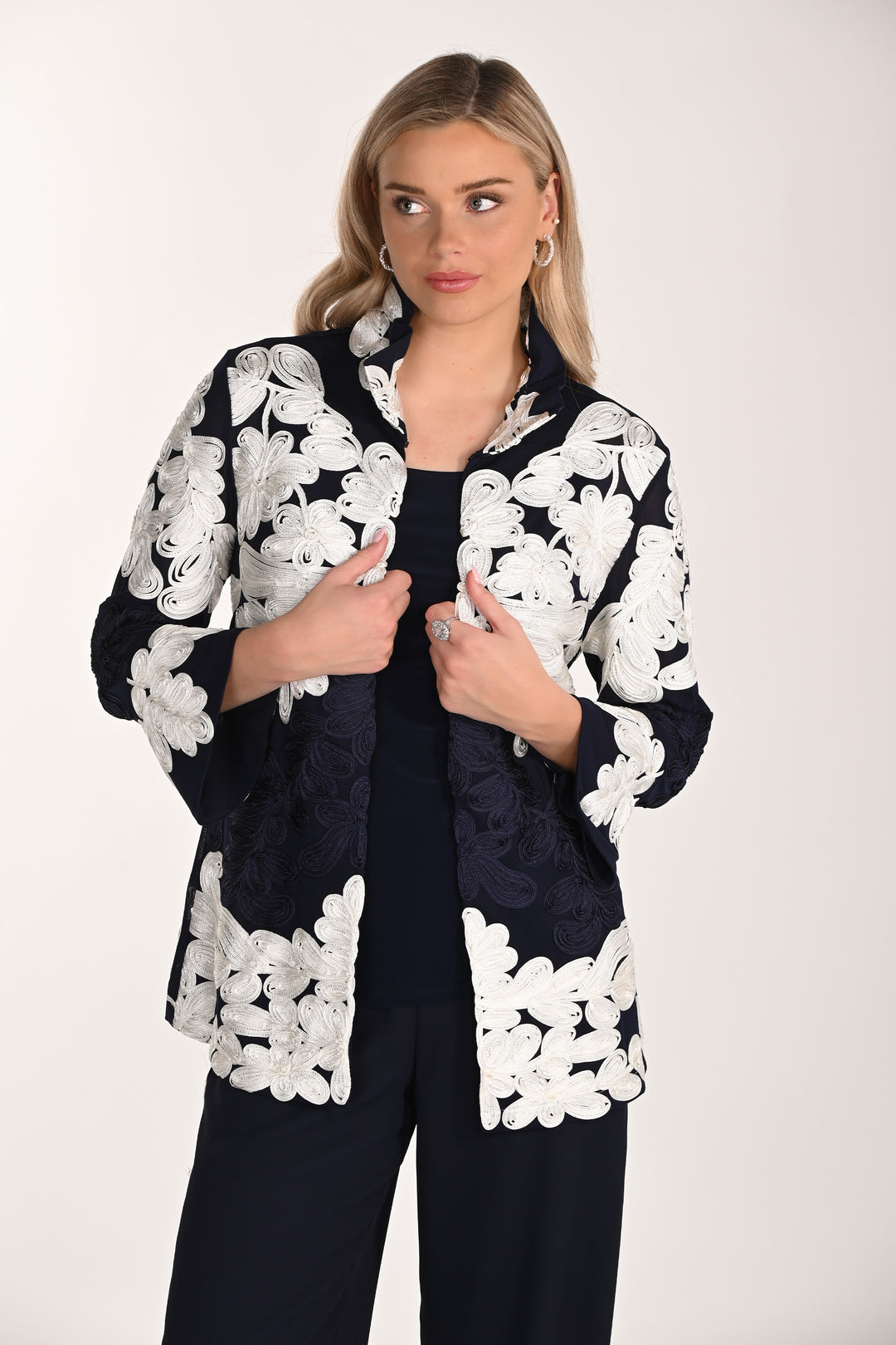 Woman wearing a black & white floral lace jacket with white floral patterns on a plain background