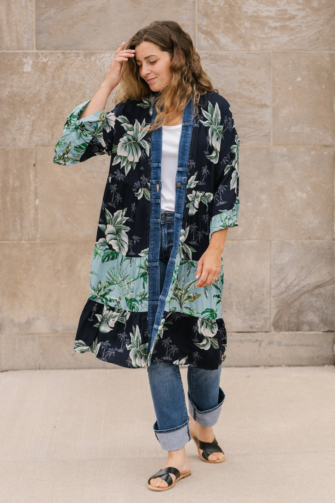 Woman wearing a floral kimono over a white shirt and jeans against a beige wall.