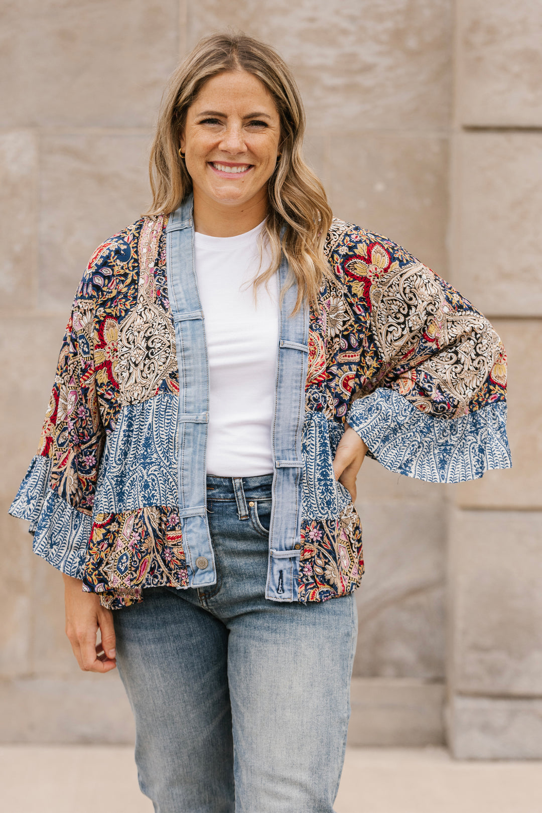 Woman wearing a colorful kimono-style jacket with ruffled sleeves against a stone wall.