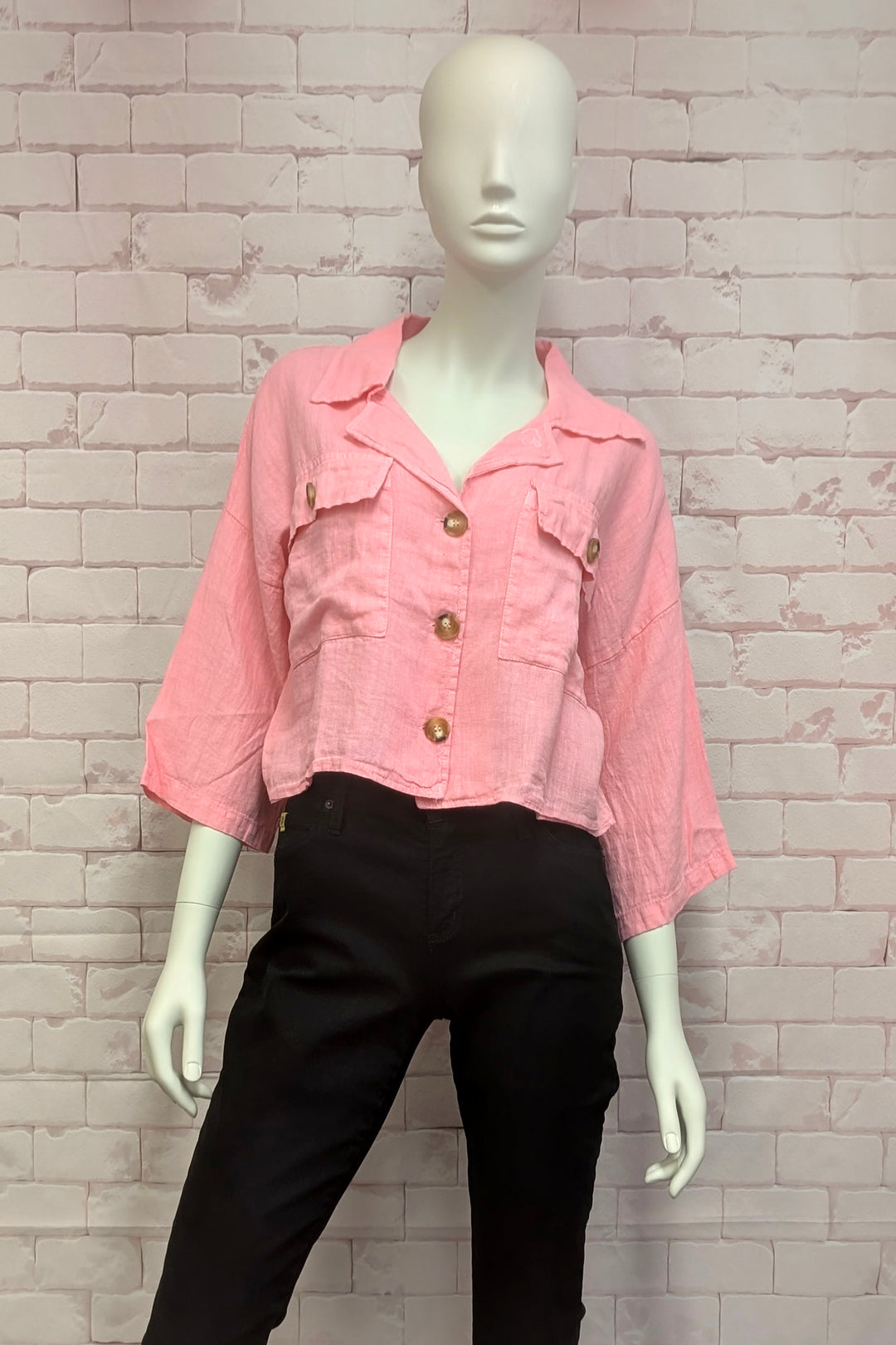 Mannequin wearing a pink linen button jacket with gold buttons against a textured white brick wall.