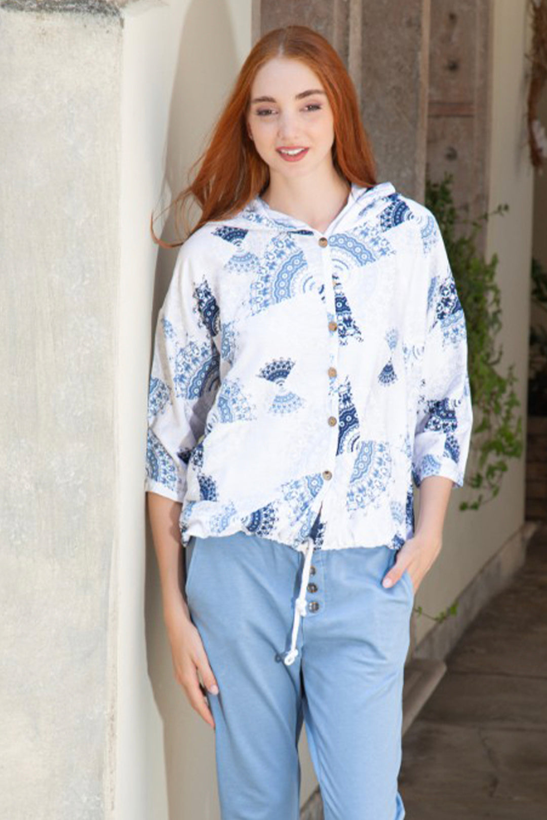 Woman wearing a white floral jacket and light blue pants standing against a textured wall.