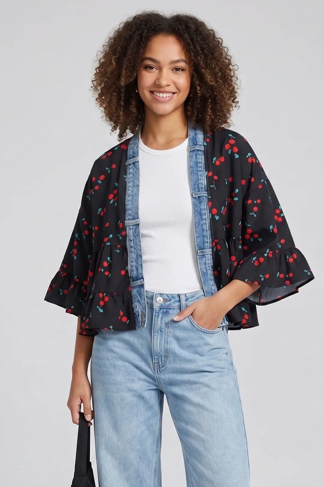 Woman wearing a black kimono-style jacket with red cherry pattern, white shirt, and blue jeans on a light gray background.