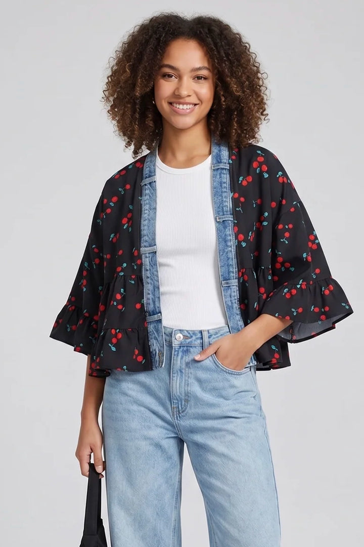Woman wearing a black kimono-style jacket with red cherry pattern, white shirt, and blue jeans on a light gray background.