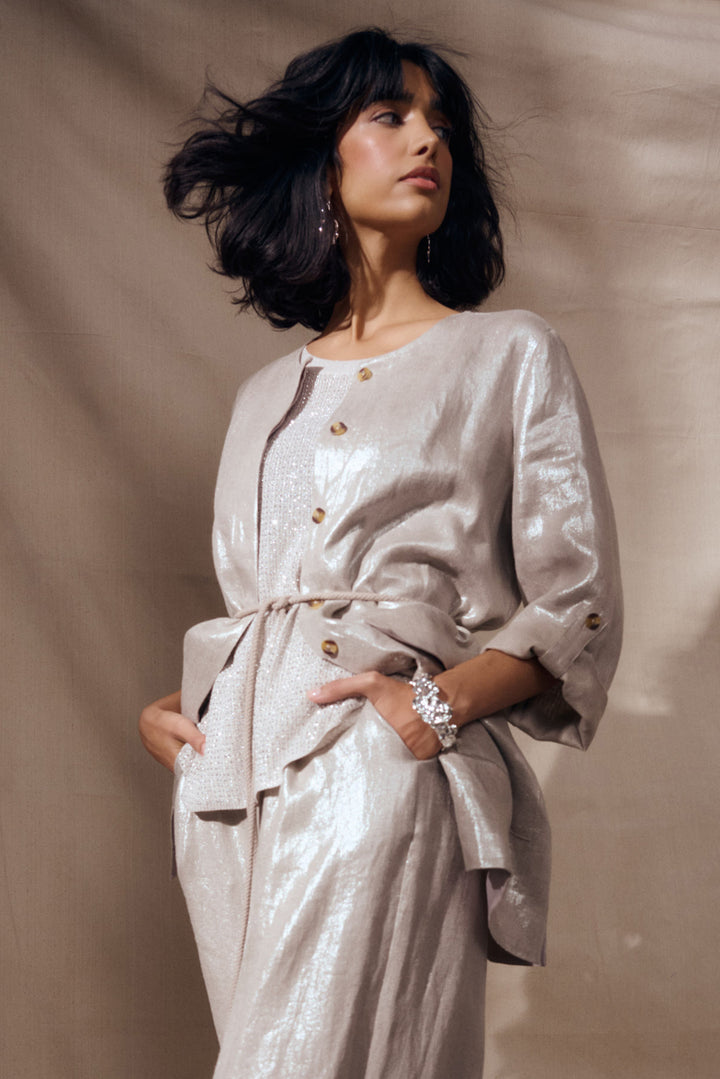 A woman wearing a shimmering champagne-colored belted jacket and matching trousers, posed against a draped beige fabric backdrop.