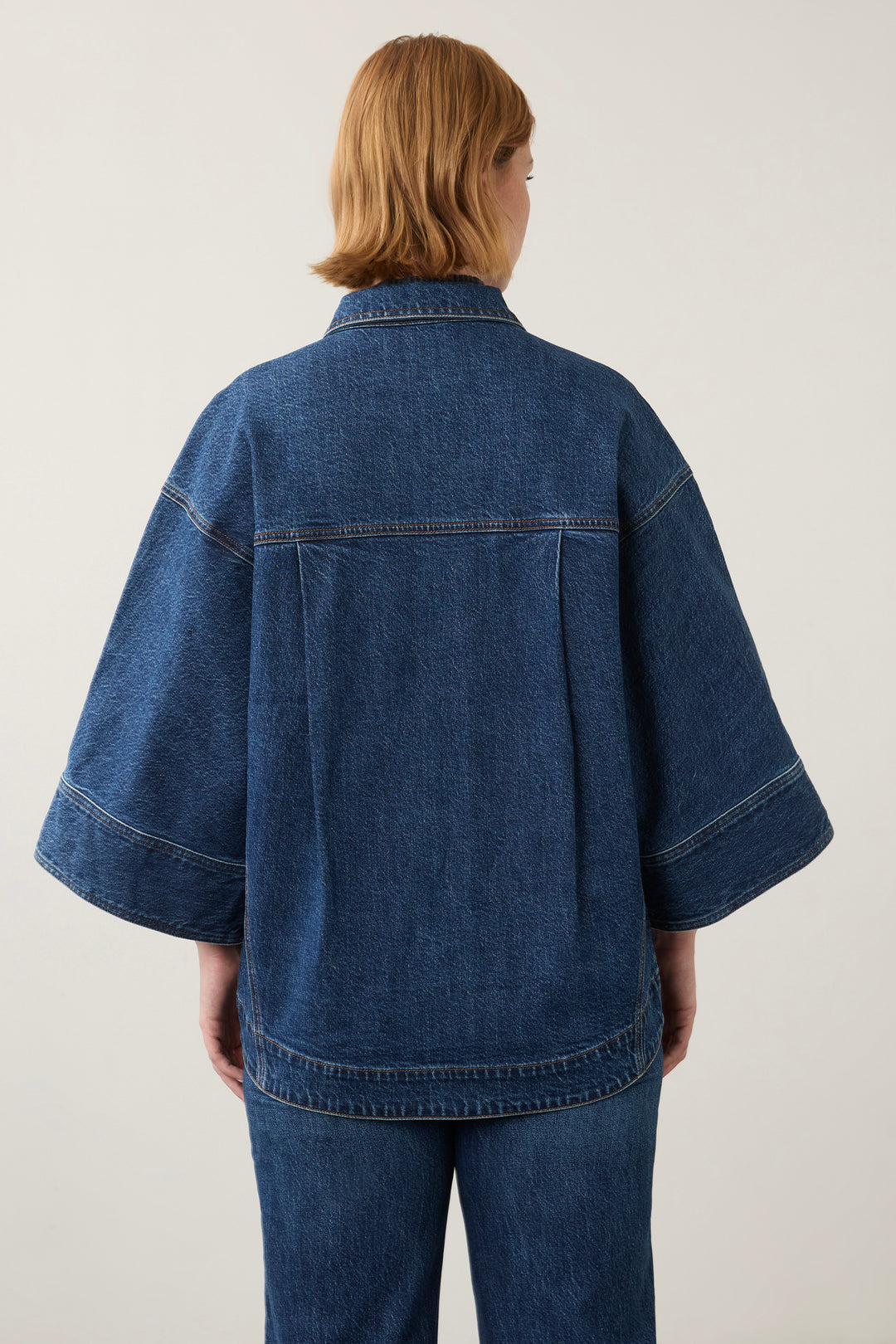 A model viewed from the back wearing a dark blue denim jacket with wide, short sleeves and matching dark blue denim pants, set against a plain light gray studio background.