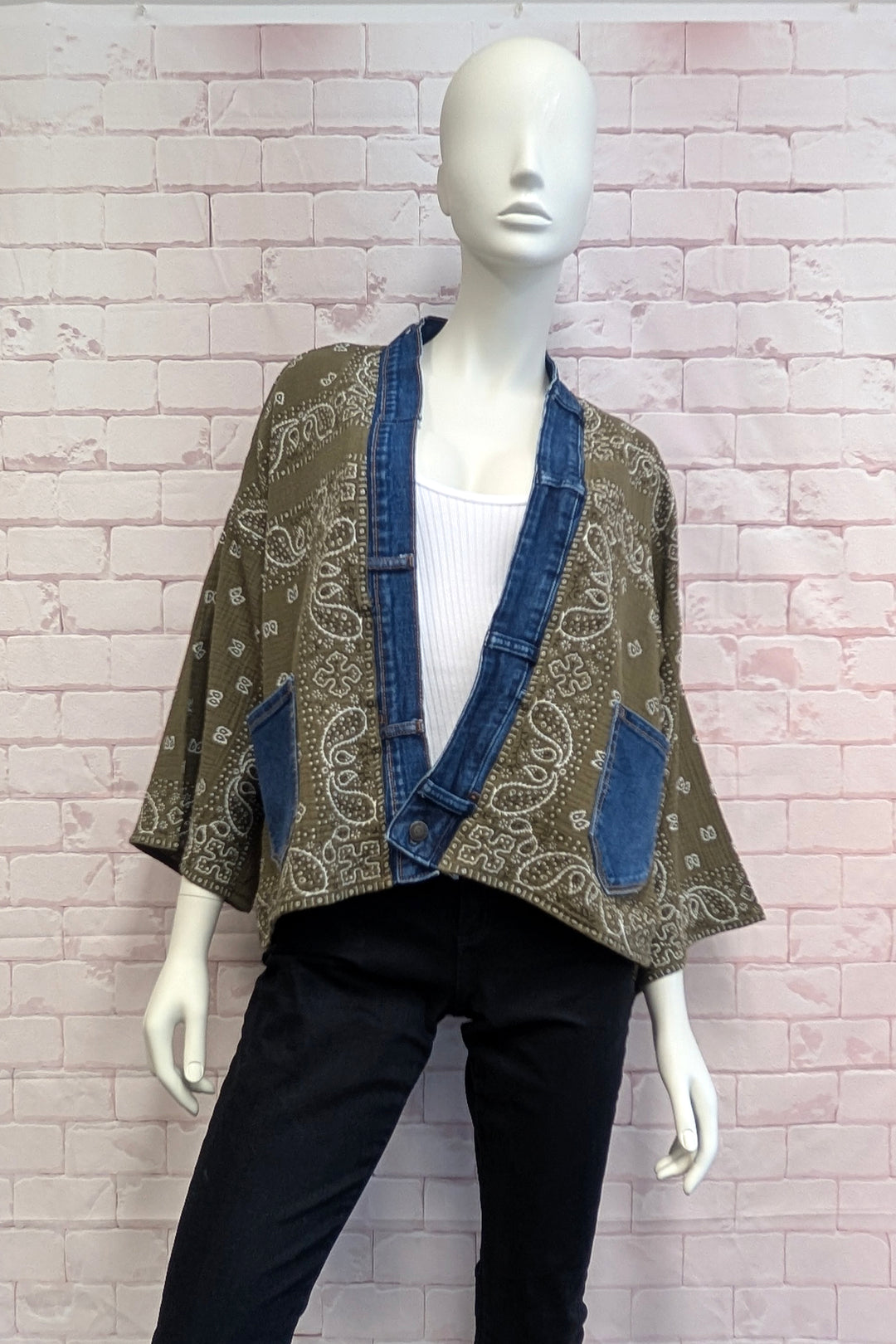 A mannequin wearing a bohemian-style open-front jacket with a brown paisley bandana print and blue denim trim, styled with a white top and black pants against a pink brick wall.