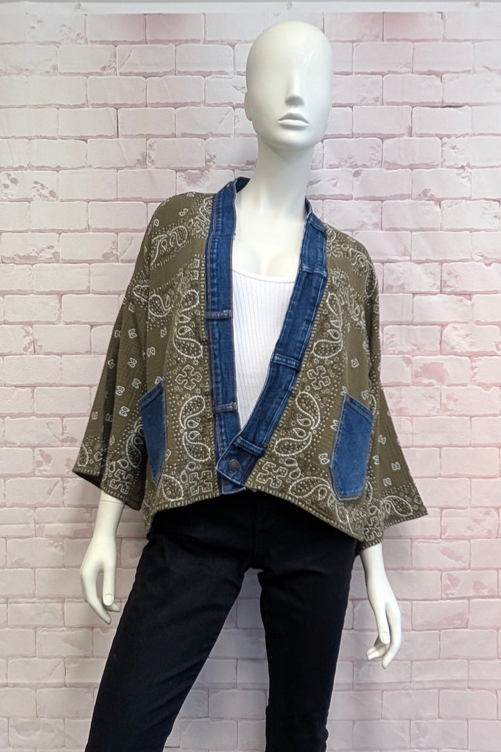A mannequin wearing a bohemian-style open-front jacket with a brown paisley bandana print and blue denim trim, styled with a white top and black pants against a pink brick wall.