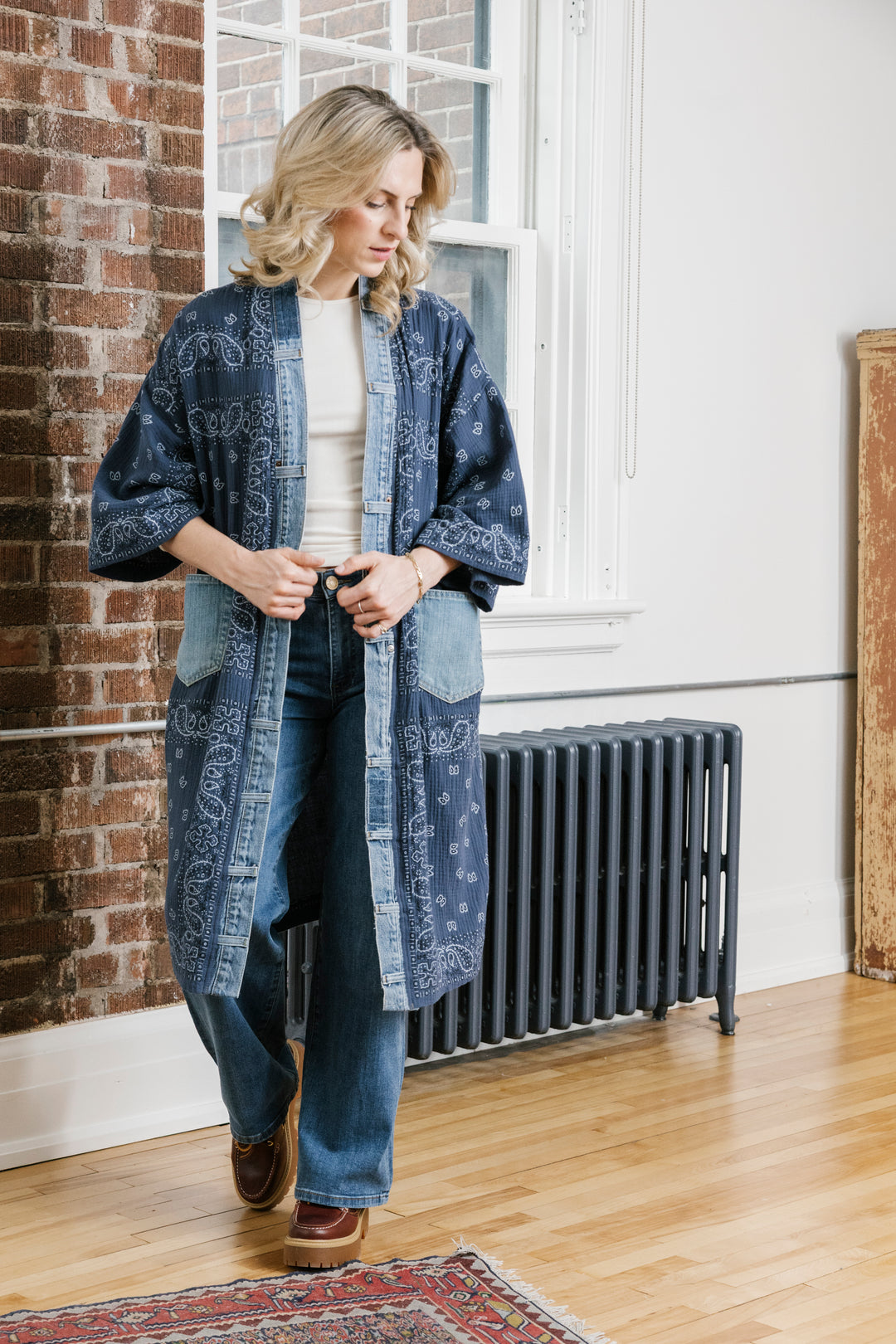 A woman wearing a blue denim kimono-style cardigan with white paisley bandana prints, styled with a white top and blue jeans, standing in a room with a brick wall, window, and wooden floor.