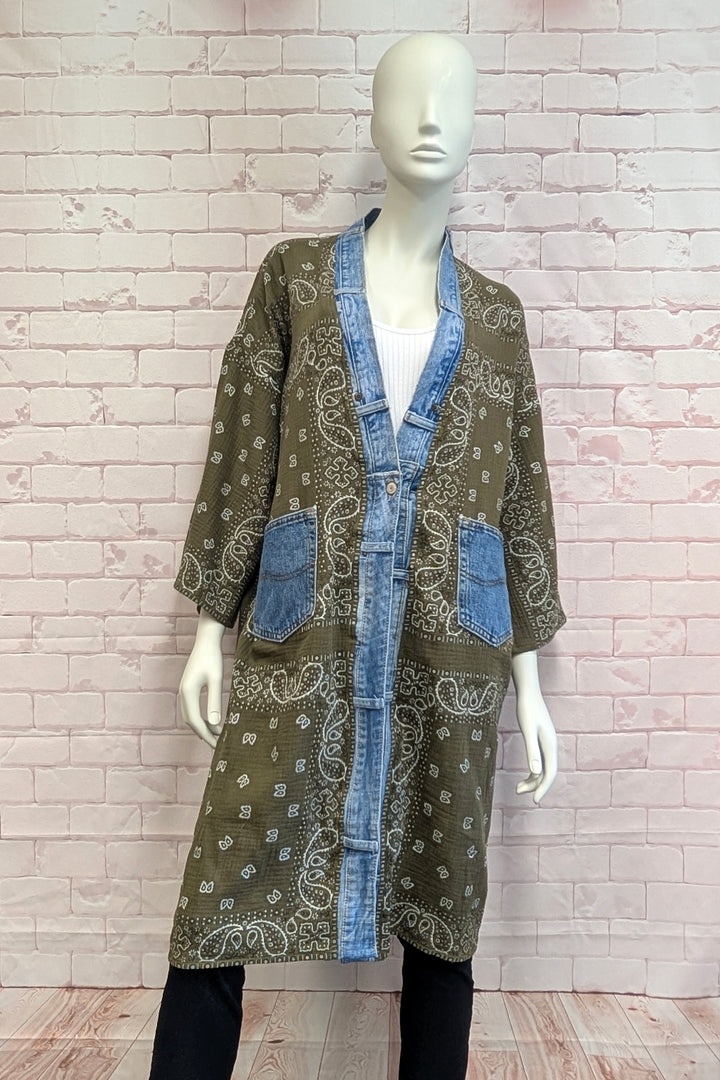 A mannequin wearing a long olive green patchwork cardigan with blue denim trim, paisley patterns, and front pockets, set against a light pink brick wall background.