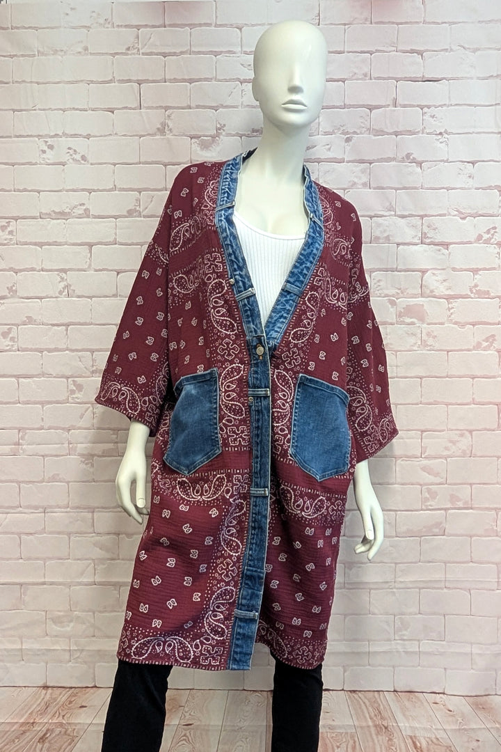 A bohemian-style open-front cardigan featuring a maroon paisley bandana print and blue denim trim, displayed on a white mannequin against a light pink brick wall.