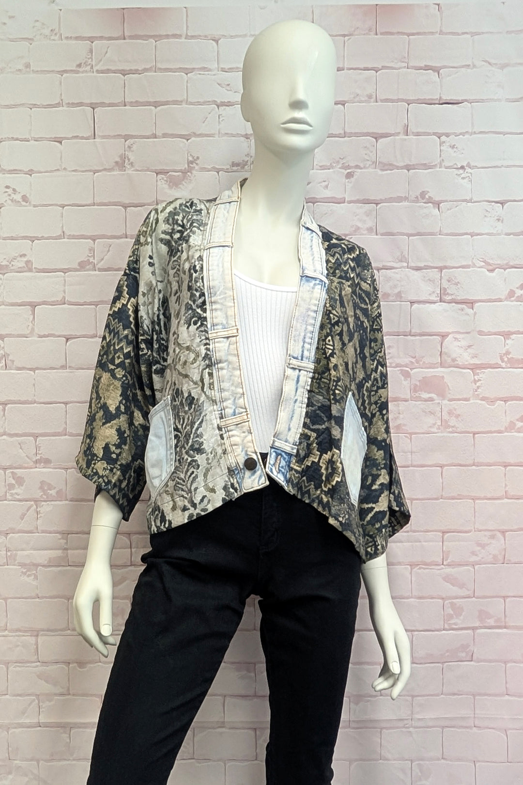 A mannequin wearing a lightweight open-front jacket with a mixed abstract print in beige, black, and gray, layered over a white ribbed tank top and black pants, set against a pink brick wall background.