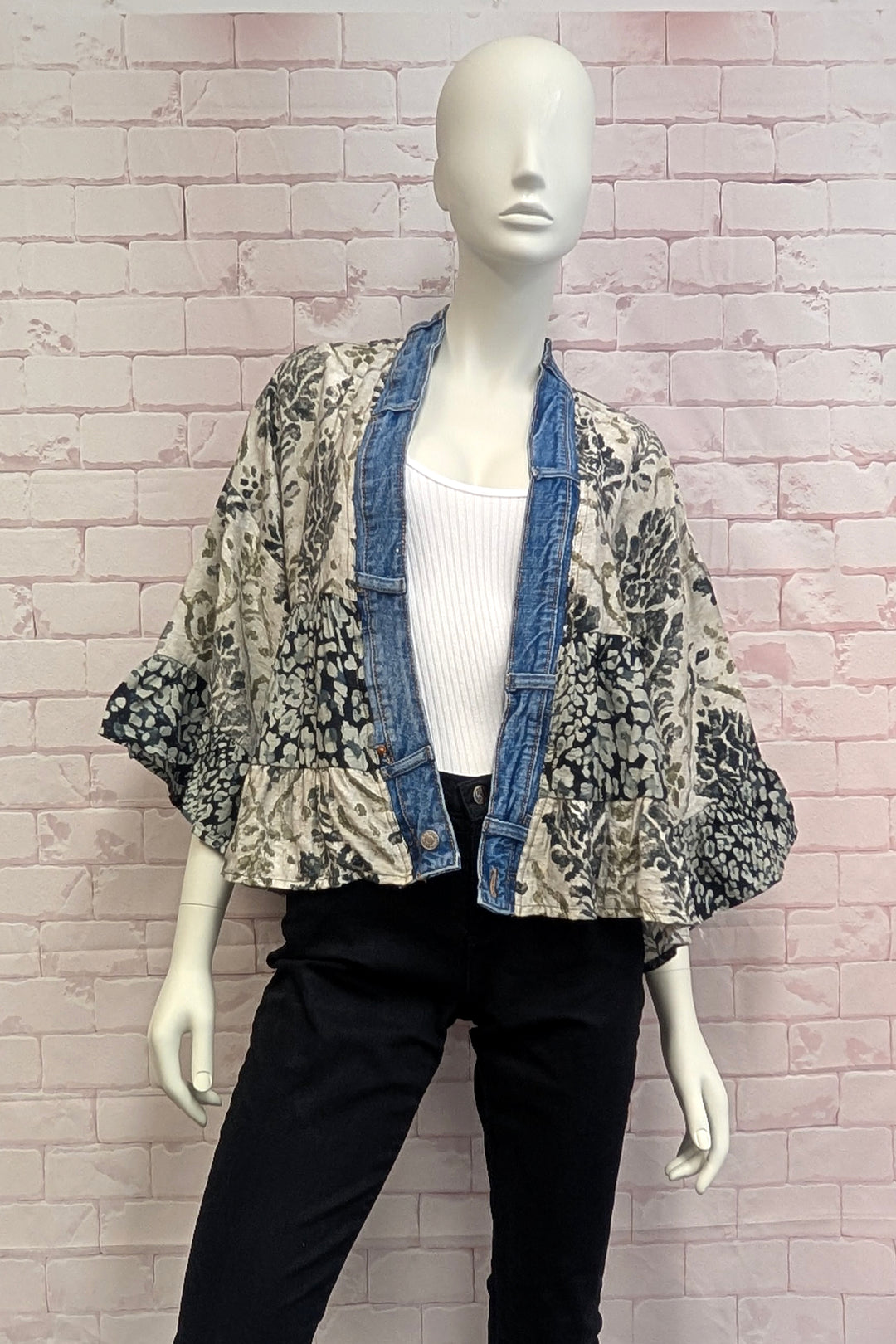 A mannequin wearing a cropped open-front jacket with a black and beige floral print over a blue denim collar, styled with a white ribbed tank top and black pants, set against a pink brick wall background.