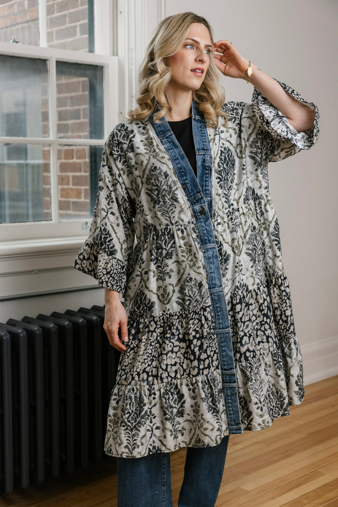 A woman models a long-sleeved, open-front kimono-style cardigan with a black and white floral ikat pattern and a blue denim button-down front, worn over a black top and blue jeans.