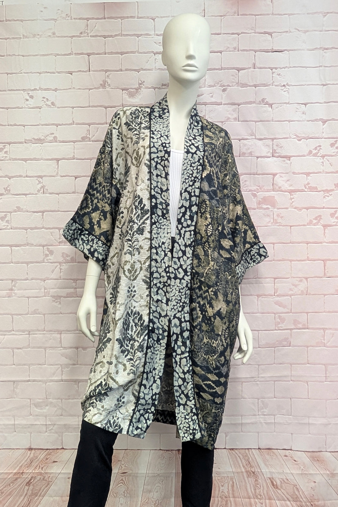 A mannequin wearing a long open-front kimono-style cardigan with a black, gray, and beige animal print, paired with black pants, set against a pink brick wall background.