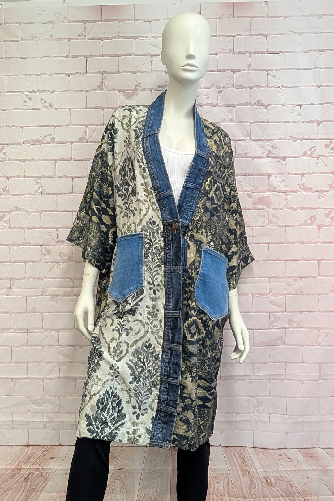 A mannequin wearing a long open-front kimono-style jacket with a mixed print pattern, blue denim trim, and two large blue patch pockets, posed against a light pink brick wall.