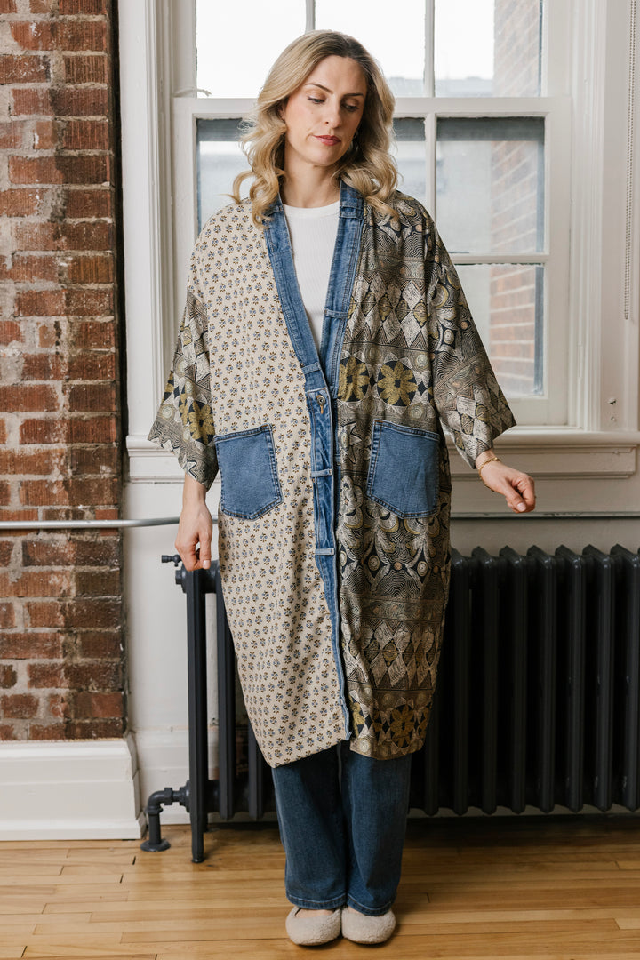 A woman stands indoors wearing a long open-front patchwork robe or kimono-style cardigan with blue denim trim and pockets, layered over a white top and blue jeans. The setting includes a brick wall, window, radiator, and wood floor.