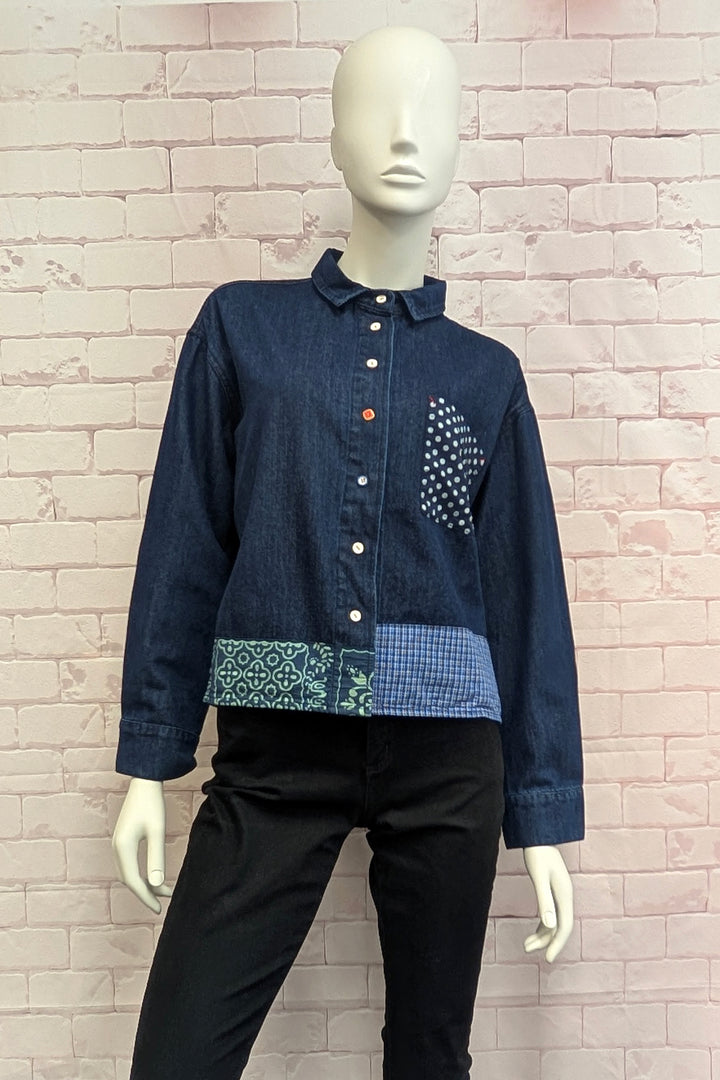 A dark blue patchwork denim shirt with patterned details, displayed on a mannequin against a light pink brick wall.