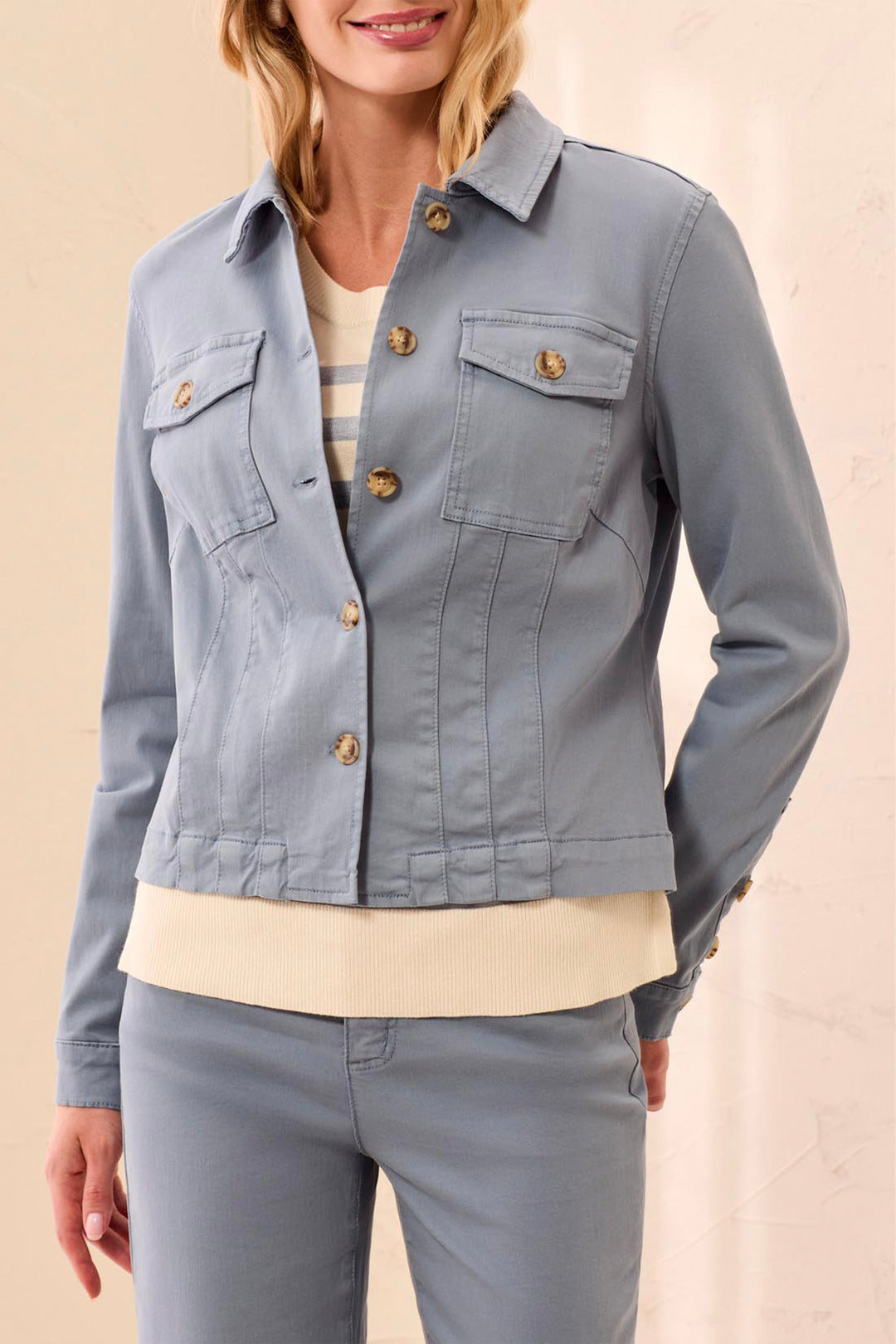 A woman wearing a light blue cropped denim jacket with gold-tone buttons over a cream top and matching blue pants, shown against a plain beige studio background.