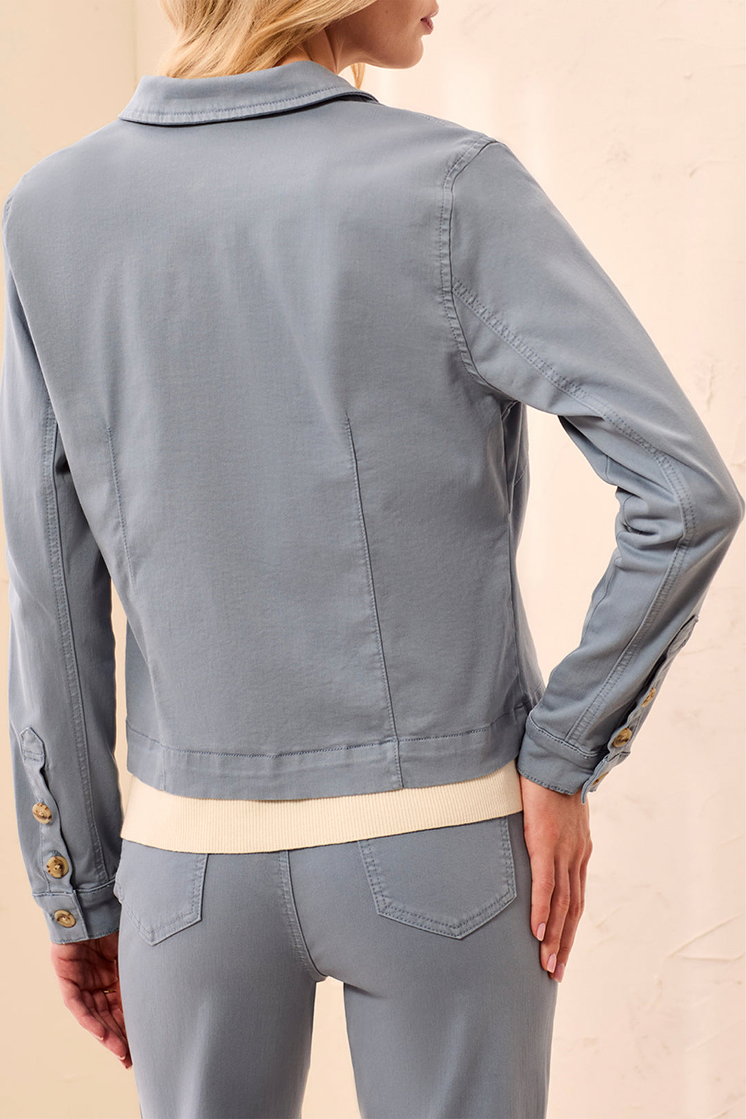 A woman shown from the back wearing a light blue-gray cropped jacket over a cream top and matching blue-gray pants, against a plain beige studio background.