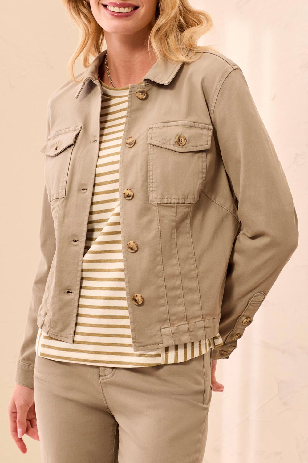 A woman wearing a light khaki utility jacket over a cream and tan striped shirt, paired with matching khaki pants, against a soft beige background.