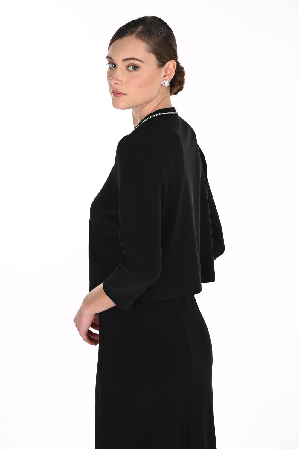 A woman wearing a black long-sleeve dress with a draped cape-style overlay, shown from a side-back angle against a plain white studio background.