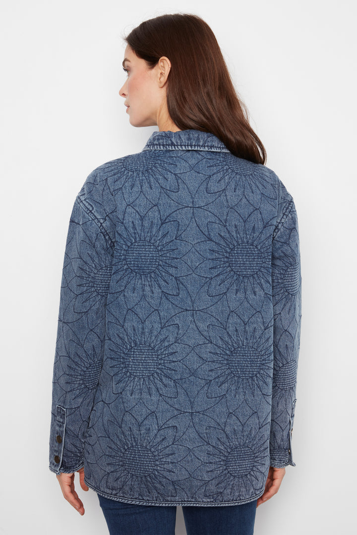 A woman shown from the back wearing a blue denim jacket with an allover raised floral sunflower pattern, paired with dark blue jeans against a plain white studio background.
