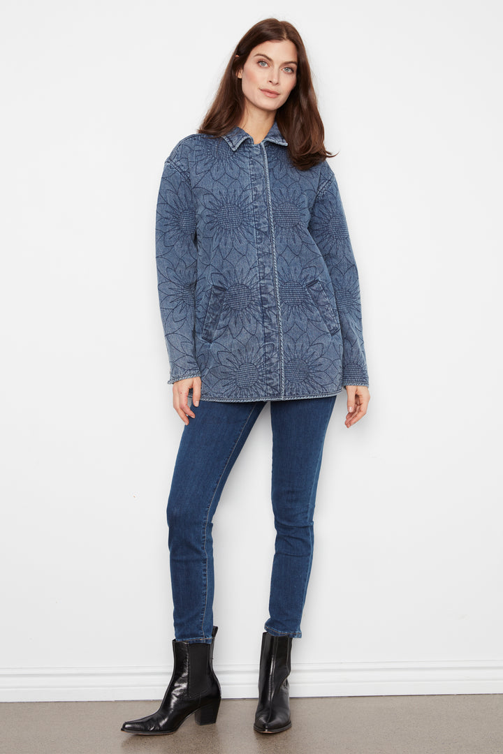 A woman modeling a blue floral jacquard button-up shirt jacket with dark blue skinny jeans and black ankle boots, standing against a white wall.