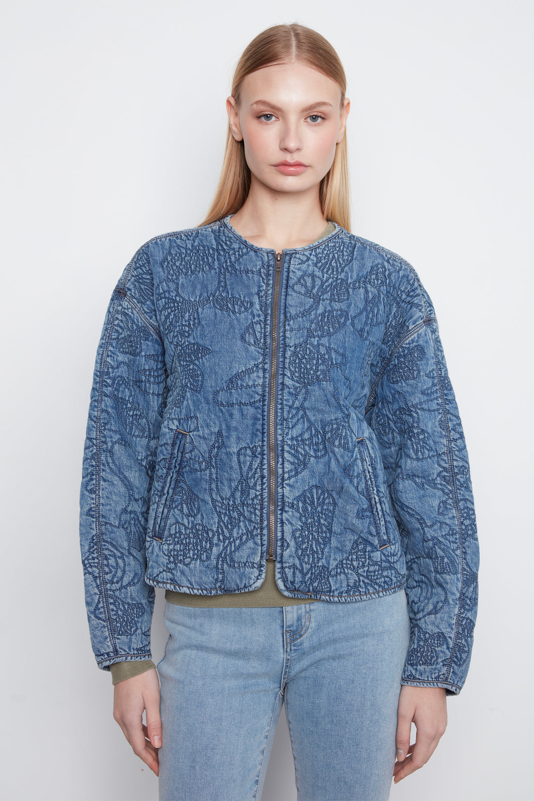 A woman modeling a cropped blue quilted jacket with a tonal floral pattern, front zipper, and zip pockets, paired with light-wash jeans against a plain light gray studio background.