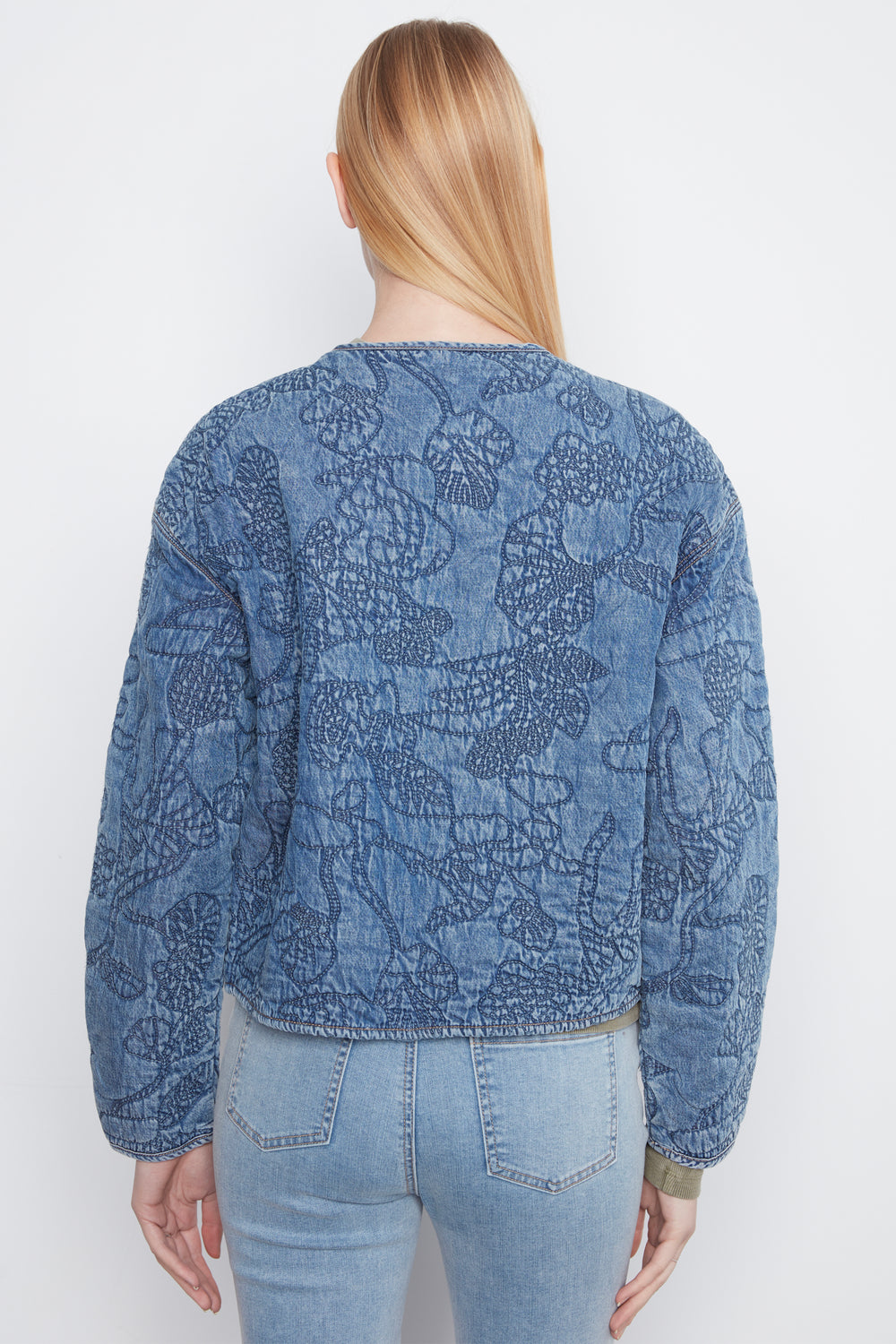 A woman viewed from the back wearing a blue long-sleeve cropped jacket with an allover tonal floral or lace-like pattern, paired with light-wash jeans against a plain light gray studio background.