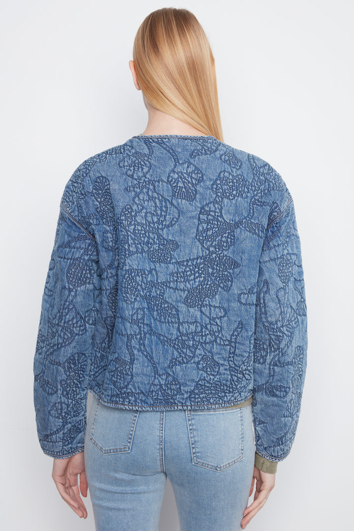 A woman viewed from the back wearing a blue long-sleeve cropped jacket with an allover tonal floral or lace-like pattern, paired with light-wash jeans against a plain light gray studio background.