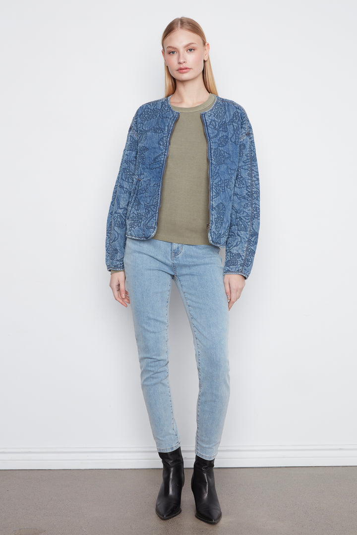 A woman modeling a cropped blue quilted jacket with a tonal floral pattern, front zipper, and zip pockets, paired with light-wash jeans against a plain light gray studio background.