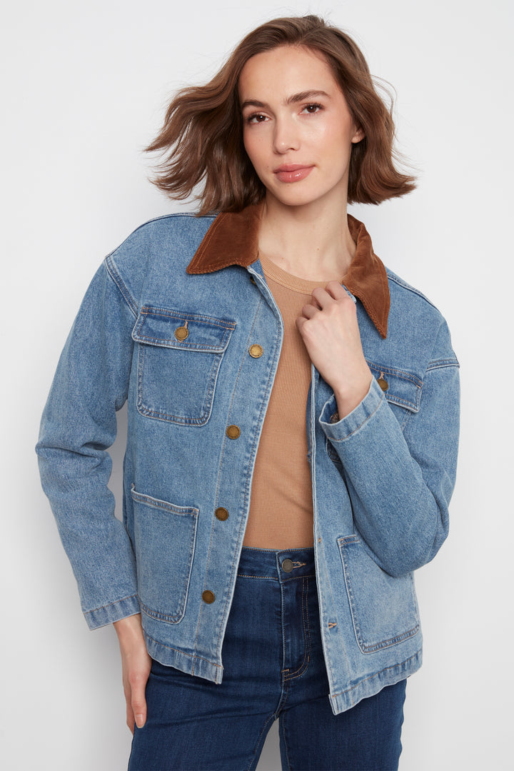 A woman modeling a light blue denim jacket with brown suede-like collar and pocket trim, paired with dark blue jeans, standing against a plain white wall.