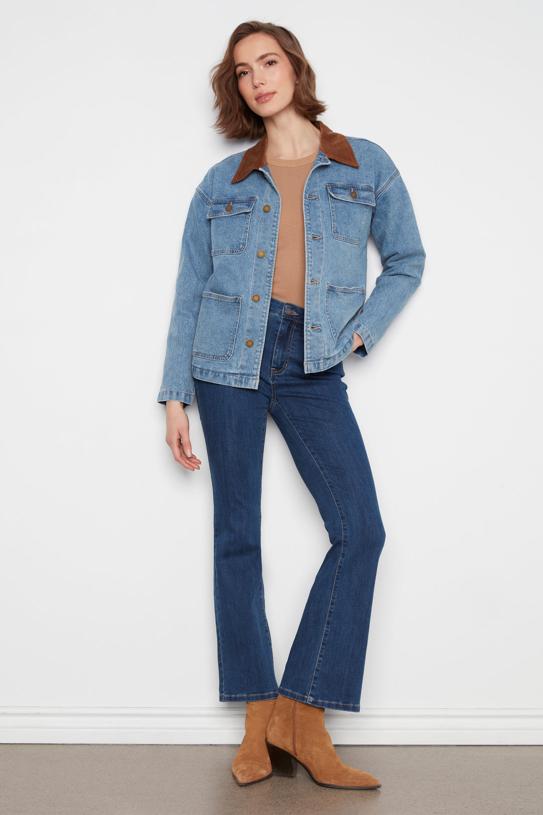 A woman modeling a light blue denim jacket with brown suede-like collar and pocket trim, paired with dark blue jeans and tan ankle boots, standing against a plain white wall.