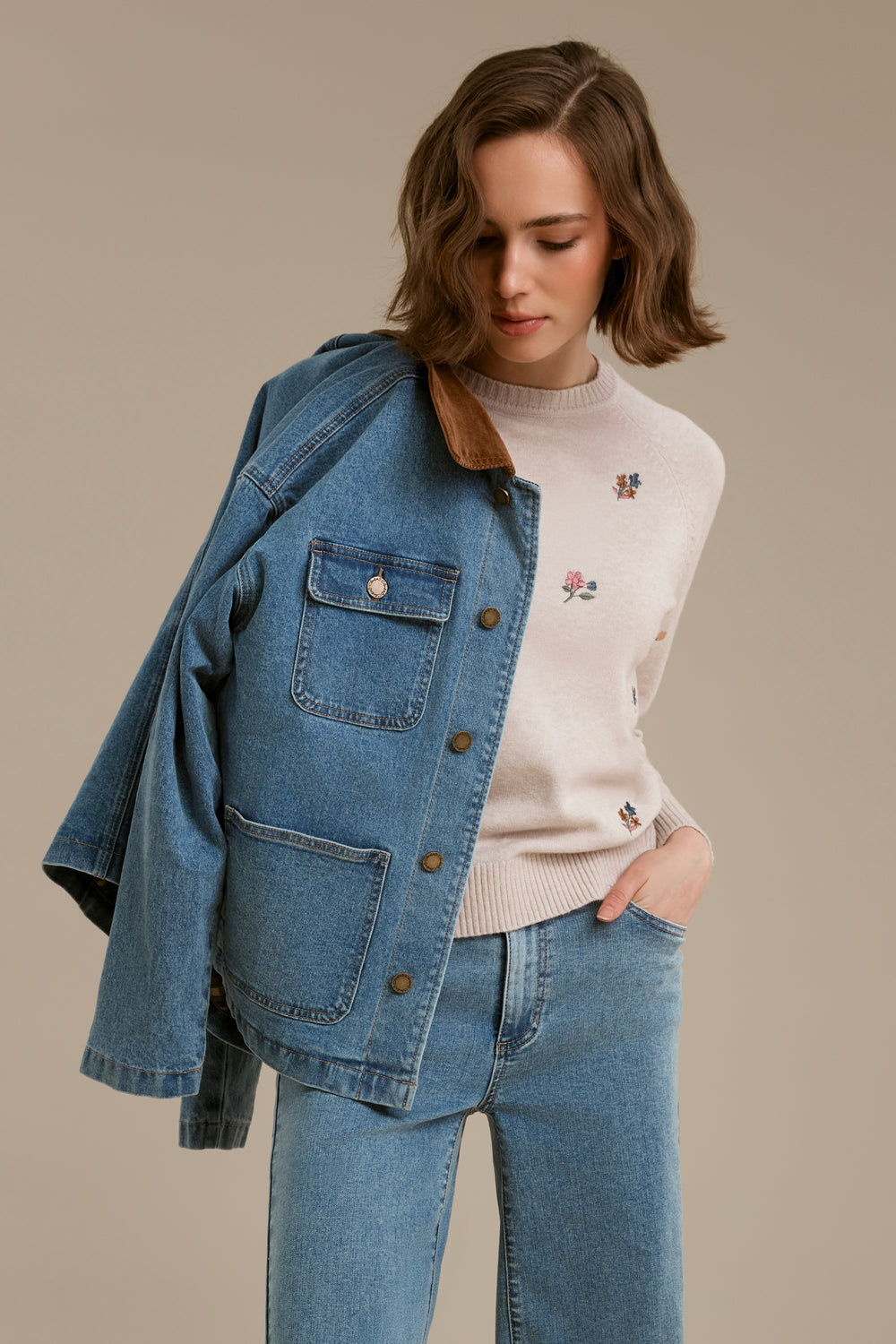A woman wearing a light blue denim jacket with brown corduroy collar, a cream ribbed sweater with floral embroidery, and light blue jeans, posed against a solid beige studio background.