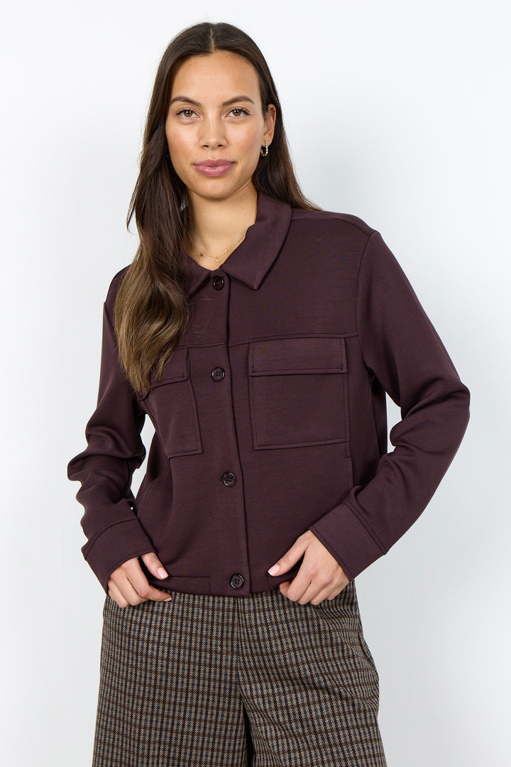 A woman modeling a dark plum button-up jacket with a pointed collar, button front, and flap chest pockets, paired with brown and black plaid trousers, against a light gray studio background.