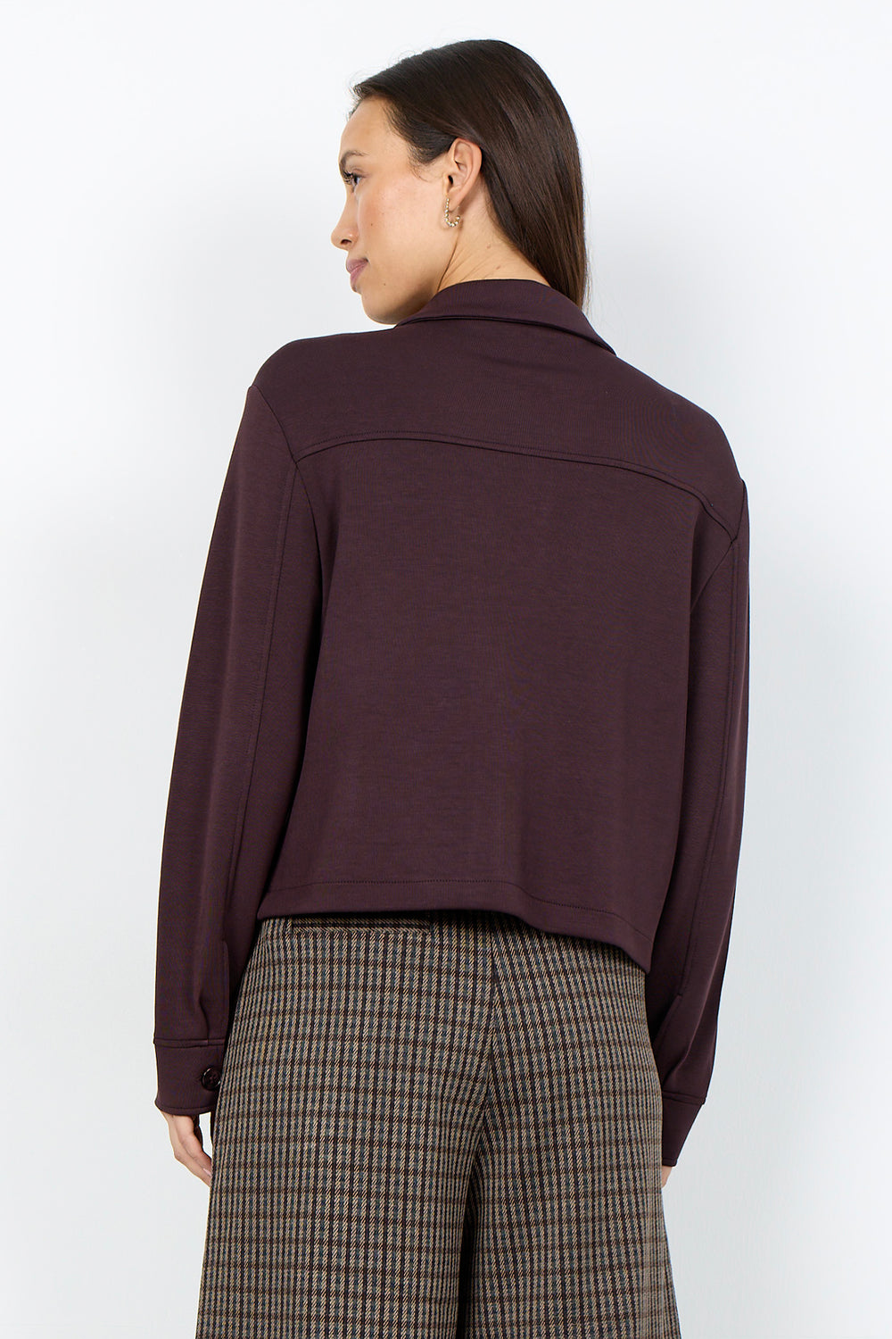 A woman shown from the back wearing a dark plum long-sleeve cropped jacket and brown-and-gray plaid trousers, photographed against a clean white studio background.